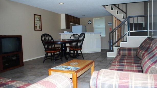 A living room with a sofa, a coffee table, and a TV. There is a kitchen area with a refrigerator and a clock on the wall.