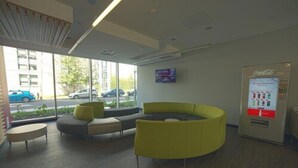 A modern waiting area with a curved yellow sofa, a flat-screen TV, and a Coca-Cola vending machine. - Queen's University - David C. Smith House - Campus Accommodation (Kingston)