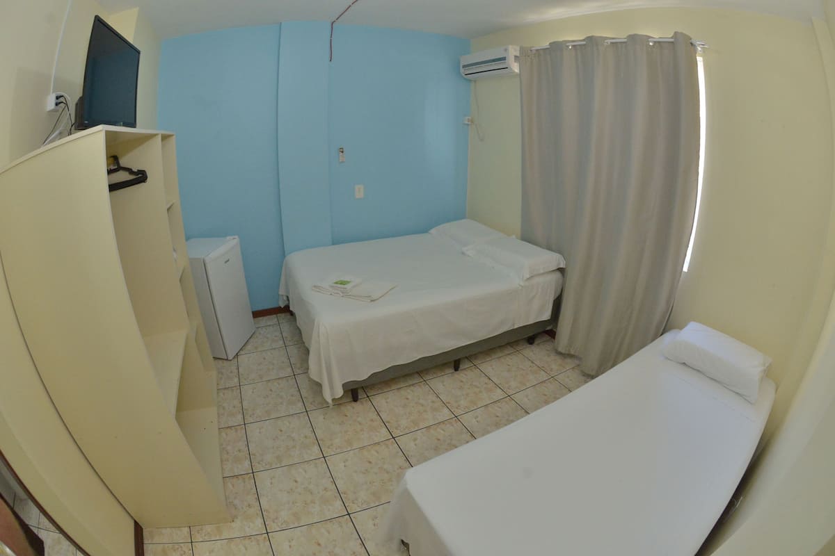 A small, single-bed room with a desk, a wardrobe, and an air conditioning unit.