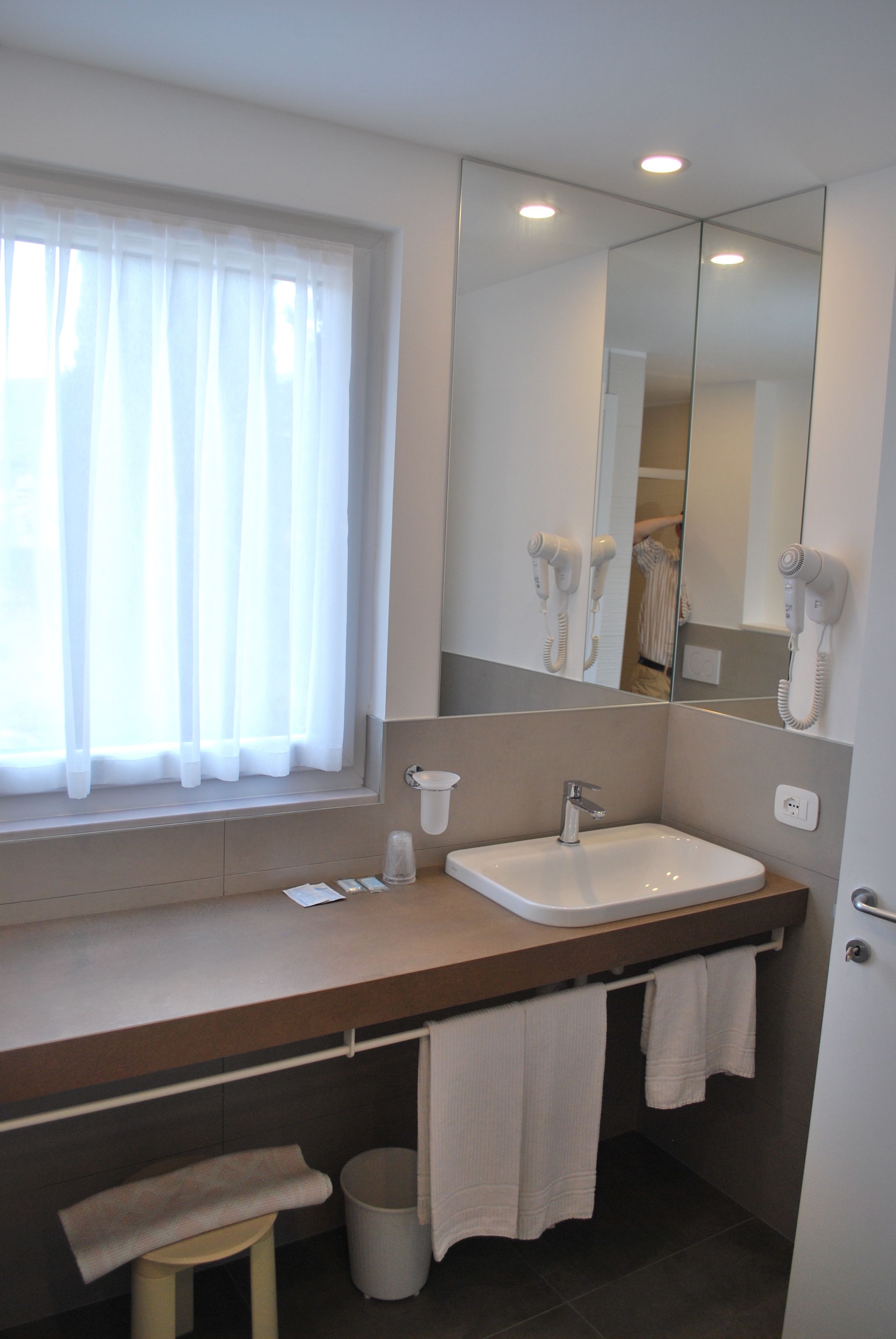 Un baño con un espejo grande, un lavamanos, una ventana con cortinas translúcidas y un perchero para toallas.