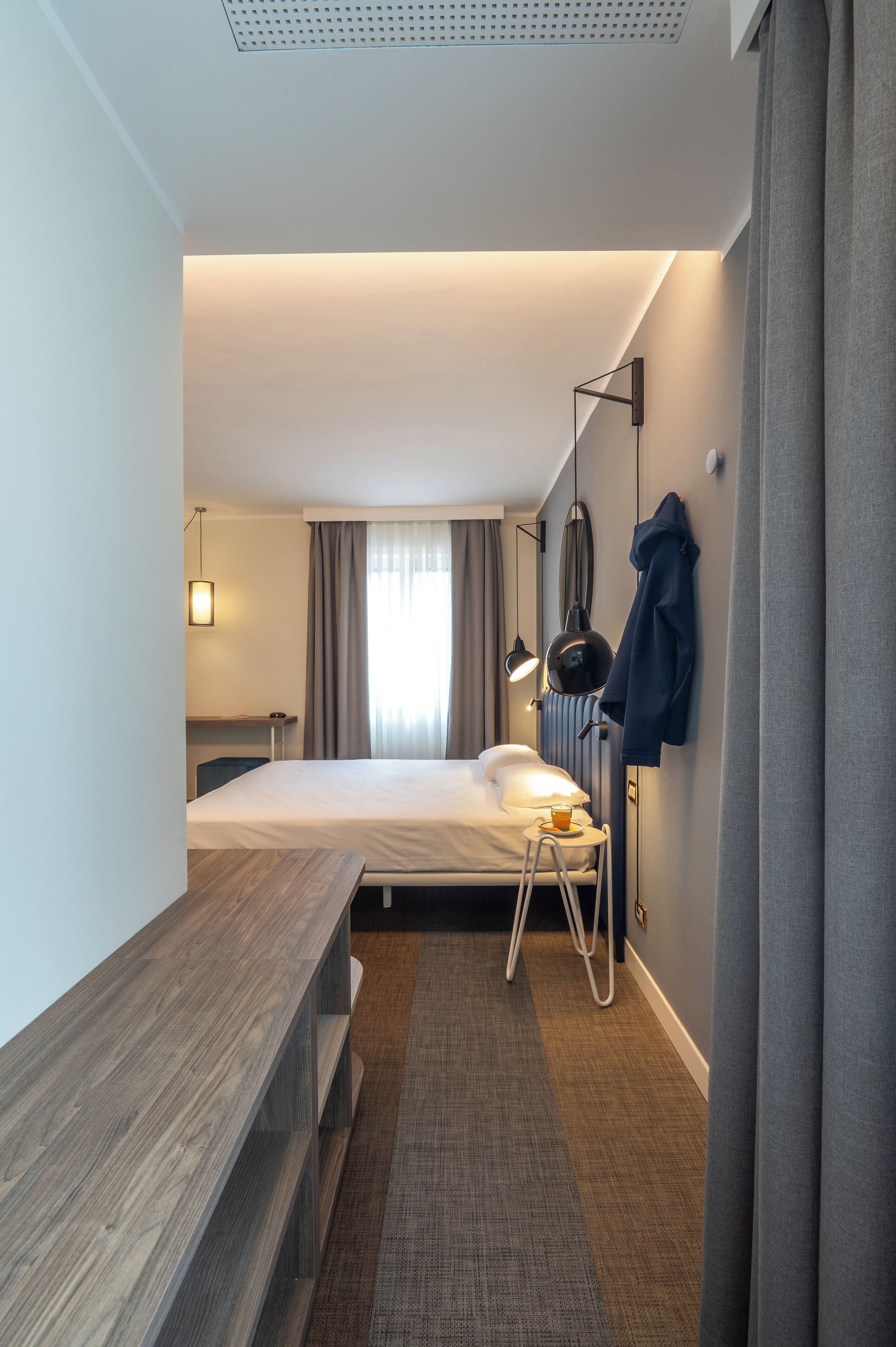 Une chambre d’hôtel de taille réduite, comprenant un lit, un bureau, une chaise, une lampe et un manteau accroché au mur.