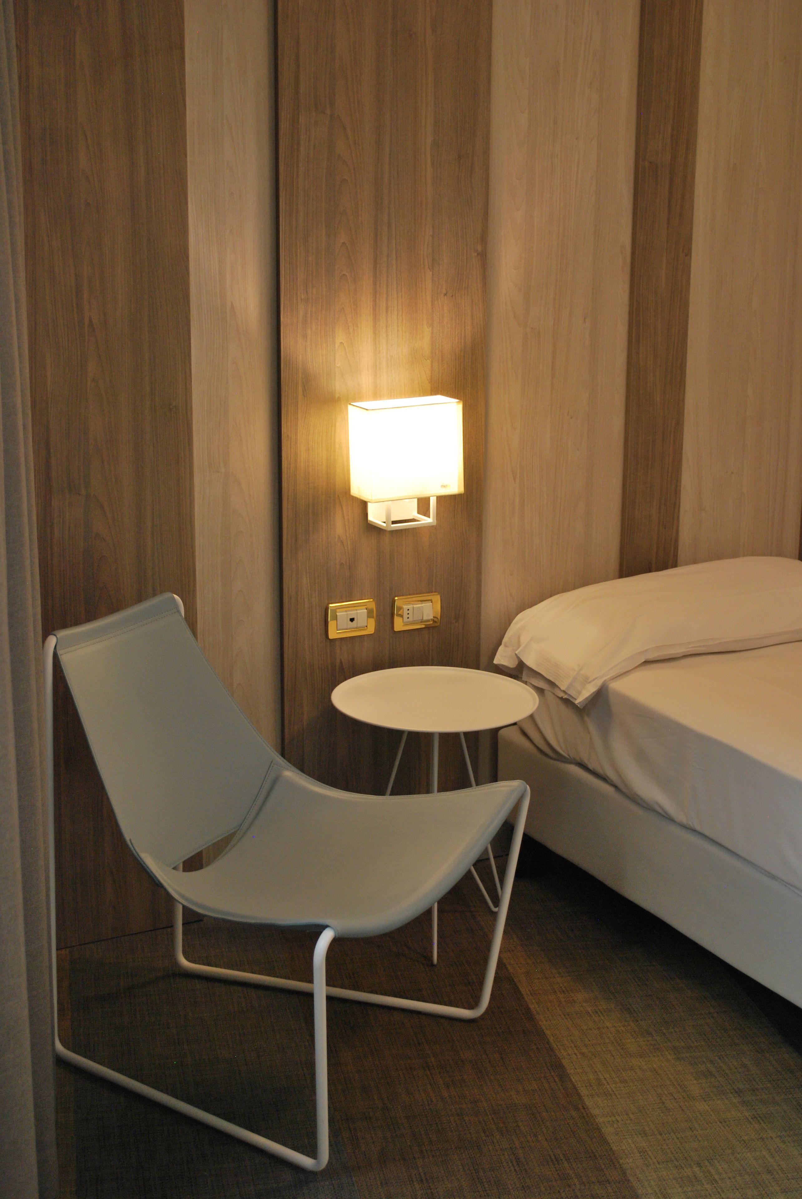 A hotel room with a single bed, a small round table, a chair, and a wall-mounted light fixture.