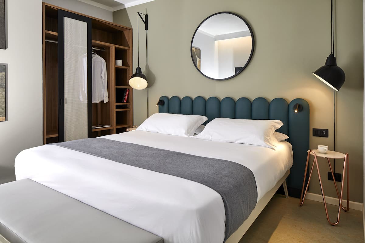 A neatly made bed with a grey and white bedspread, a round mirror on the wall, and a small table with a cup beside it.