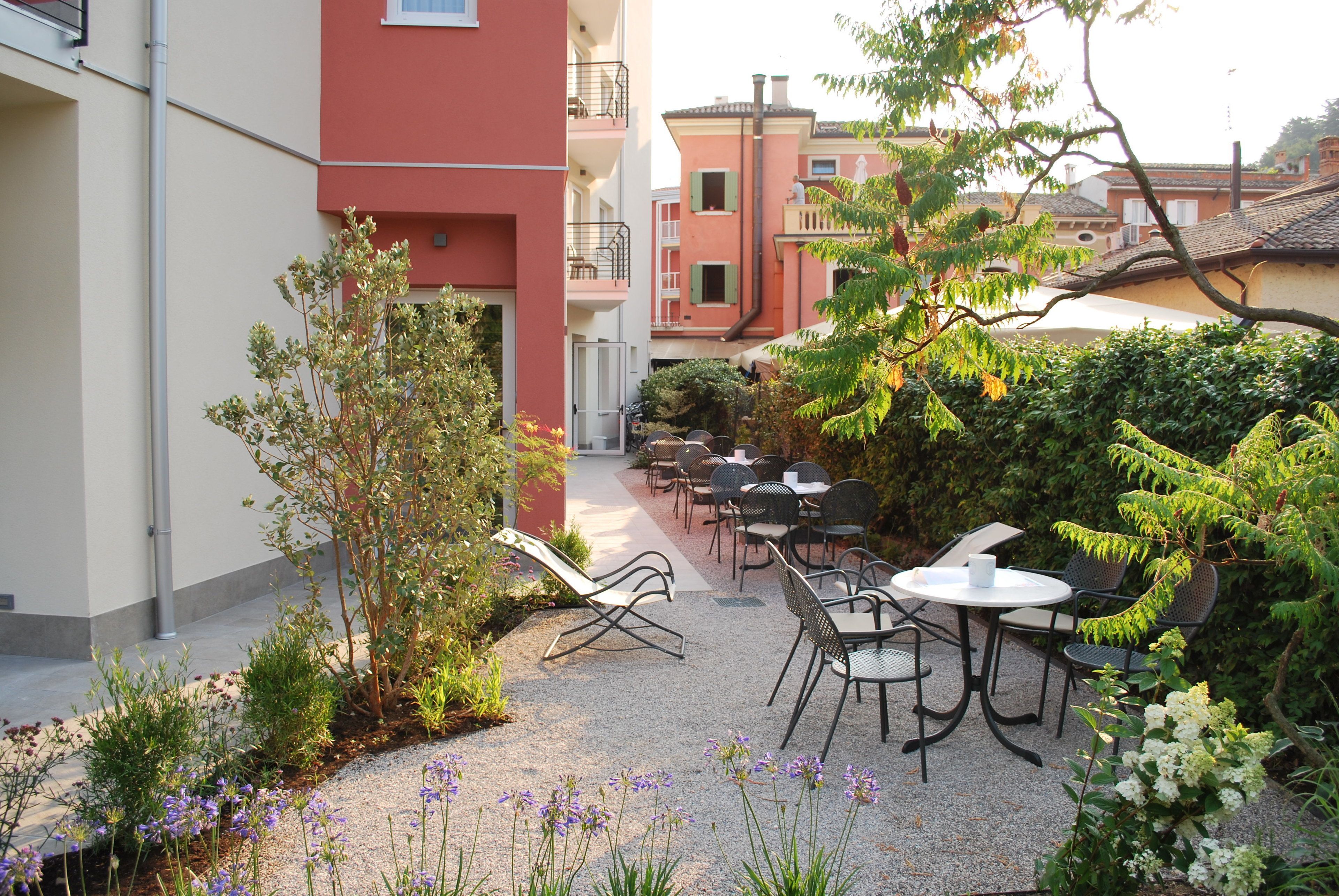 Un patio al aire libre con mesas y sillas, rodeado de follaje y un edificio al fondo.