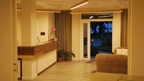 A modern living room with a glass coffee table, a sofa, and a wooden reception desk. There is a potted plant and a vase with flowers on the desk. - Hotel Europa (Garda)