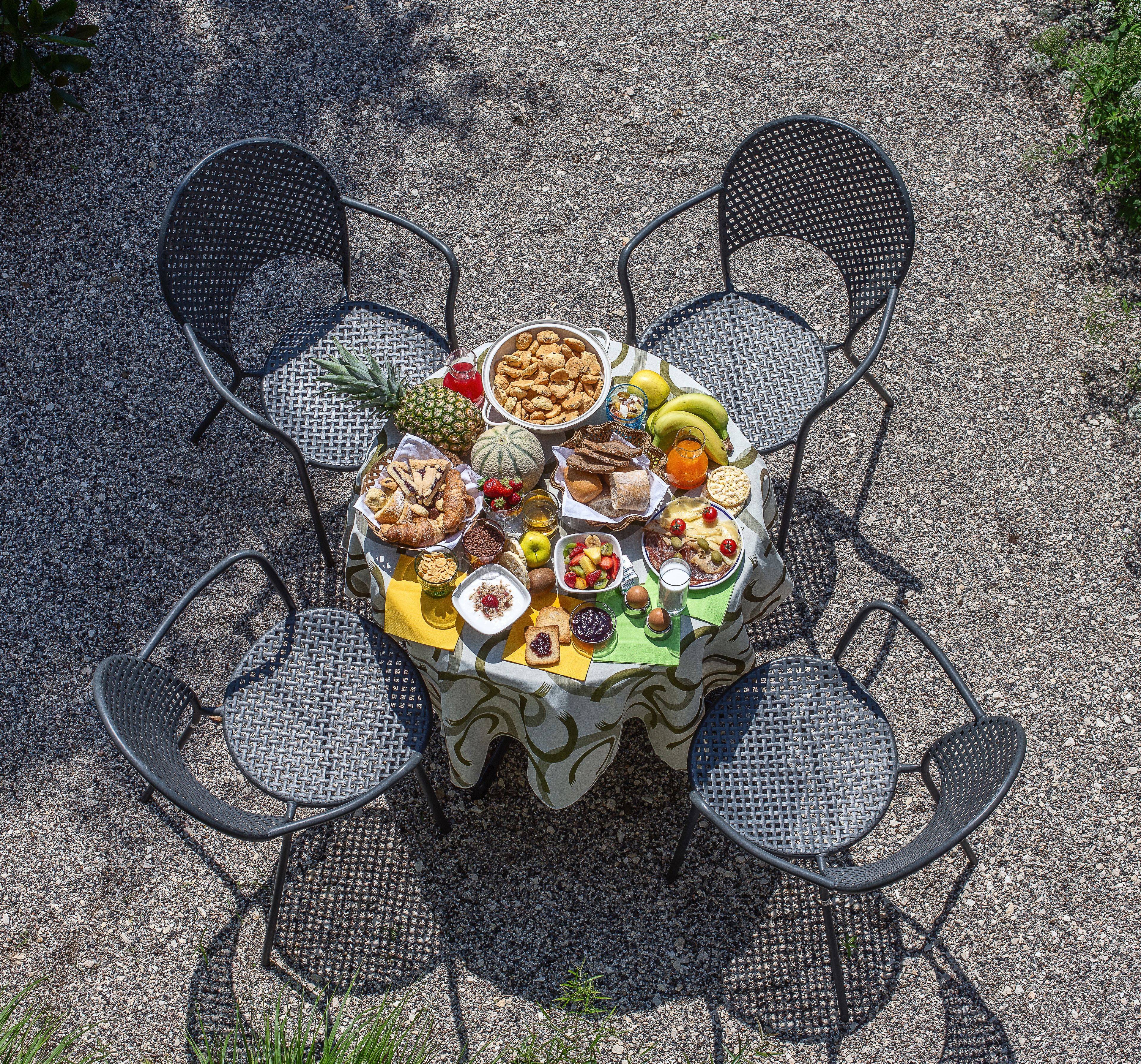 A round outdoor table with a woven tablecloth, surrounded by four black metal chairs. The table is set with various food items and drinks.