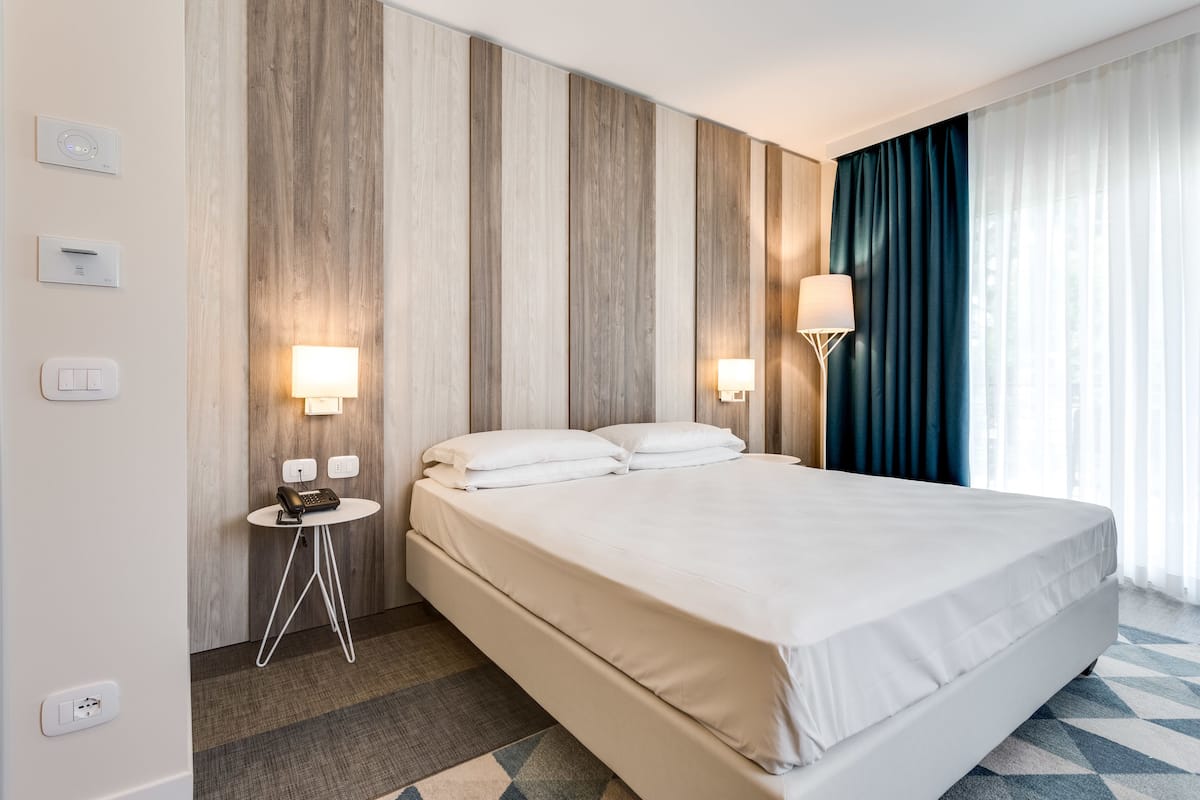 A hotel room with a large bed, two bedside tables with lamps, a wall-mounted thermostat, and a window with curtains.