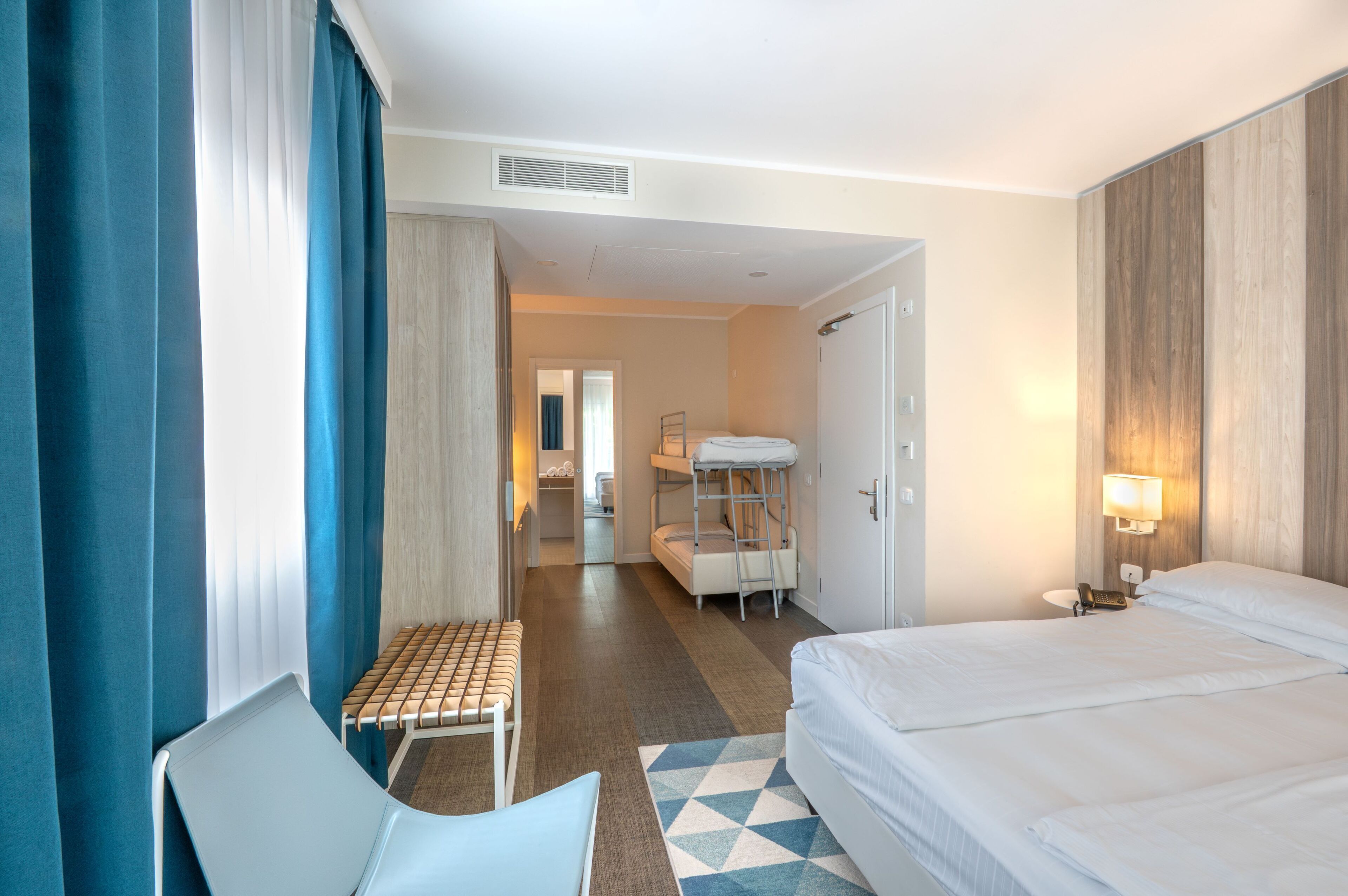 Habitación de hotel con una cama, una silla, una mesita y una cama nido al fondo.