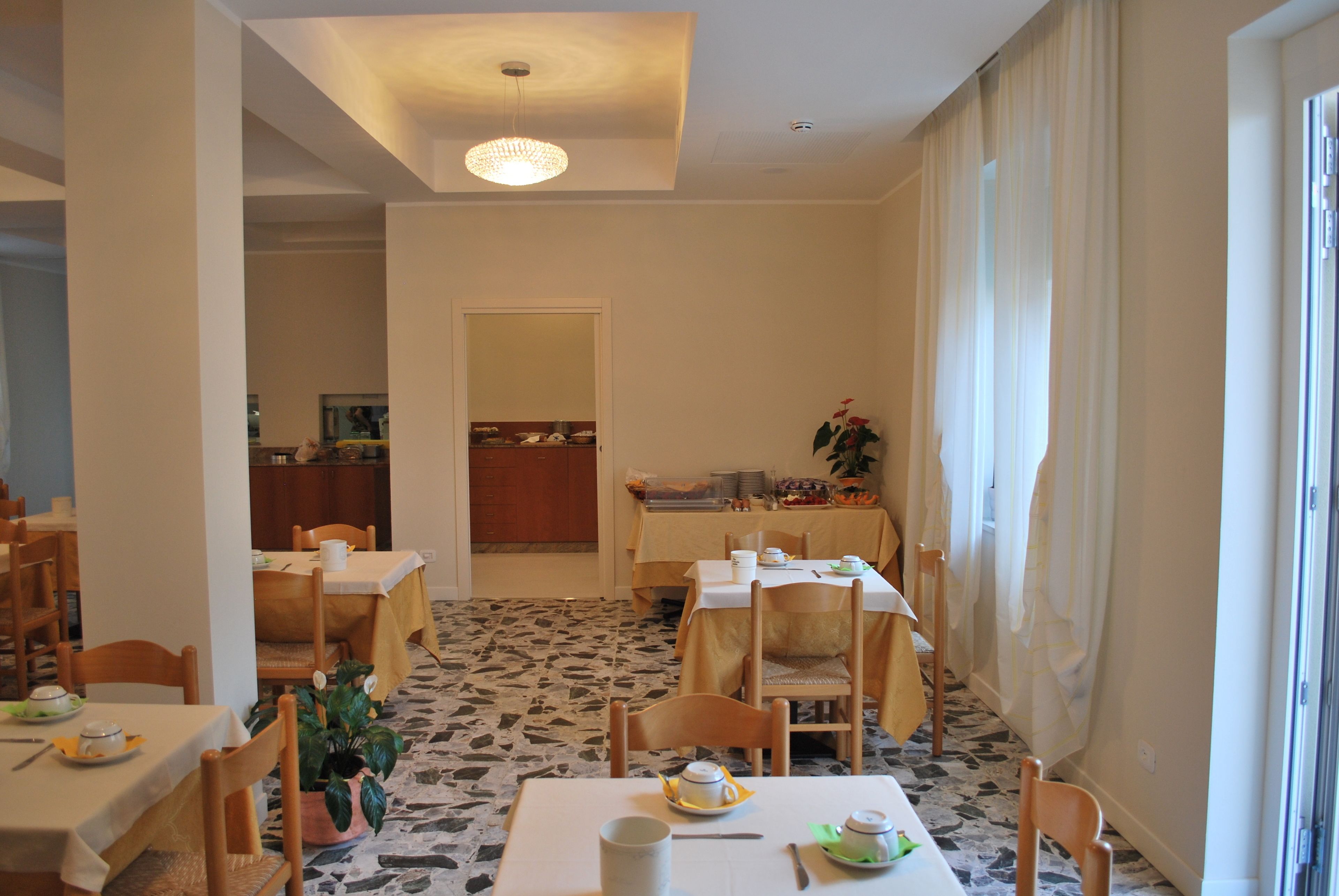 A dining area with multiple tables and chairs, a chandelier, and a view of the kitchen in the background.