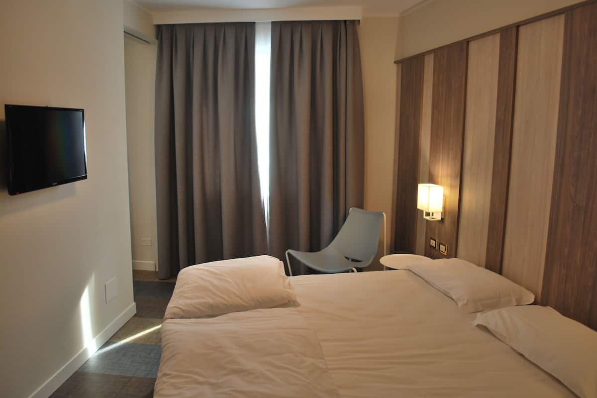 A hotel room with a large bed, a flat-screen TV mounted on the wall, a chair, and a bedside table with a lamp.