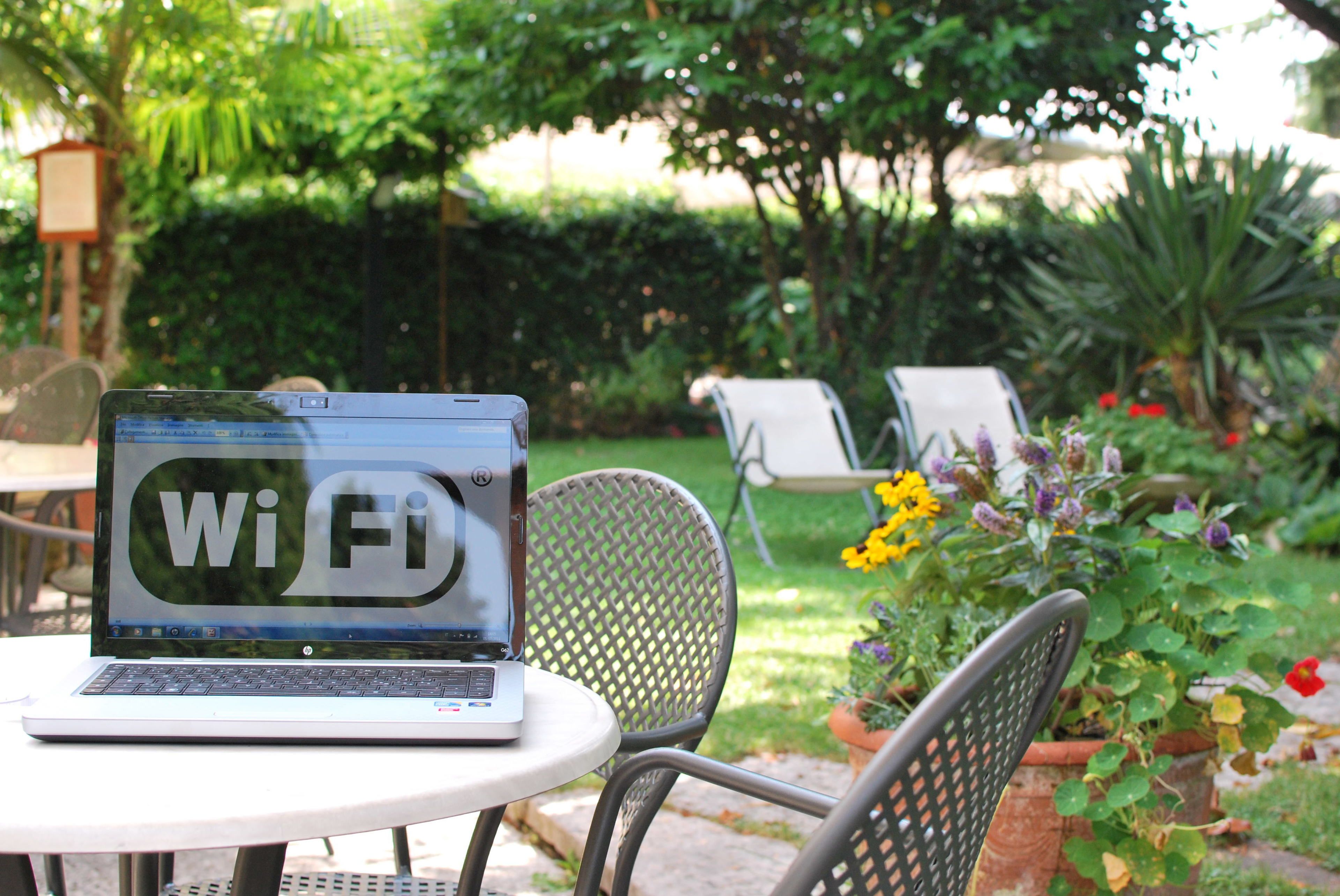 Una laptop con el símbolo de Wi-Fi en la pantalla, sobre una mesa redonda en un espacio exterior con sillas y plantas en macetas.