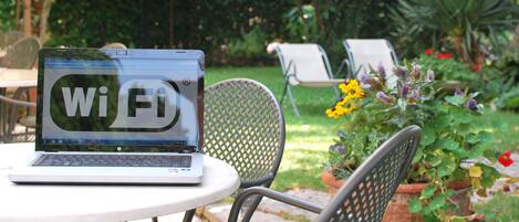 Una laptop con el símbolo de Wi-Fi en la pantalla, sobre una mesa redonda en un espacio exterior con sillas y plantas en macetas.