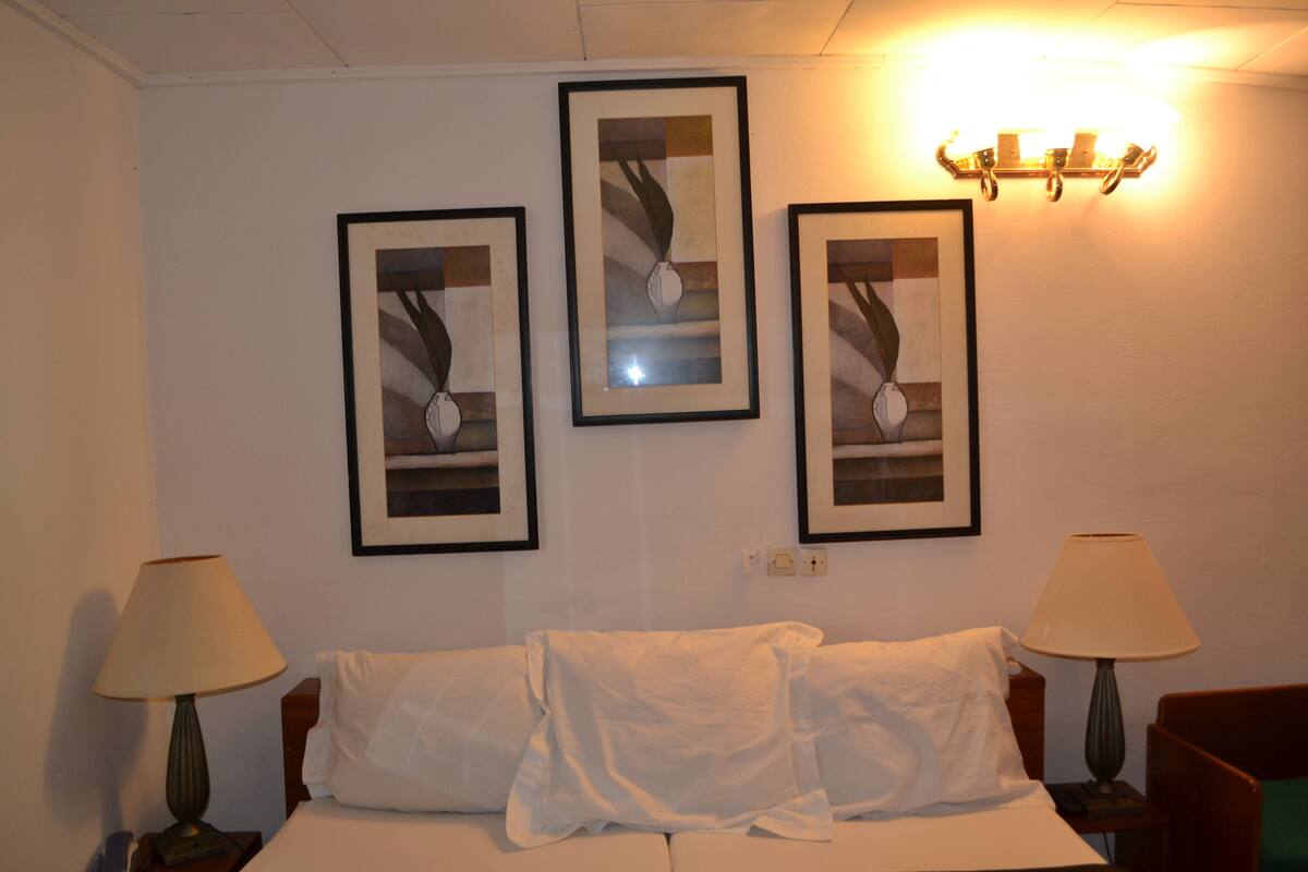 A hotel room with a bed, two bedside lamps, and three framed artworks on the wall.