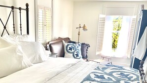 Quarto com cama grande, cabeceira de madeira, mesa de cabeceira, abajur e janela com persianas.