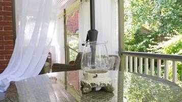 Una mesa de vidrio con una lámpara de estilo antiguo en un balcón con mobiliario de mimbre y una cortina blanca.