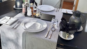 Uma mesa de jantar posta com pratos, talheres, copos e uma chaleira, com toalhas de mesa listradas e um abajur ao fundo.
