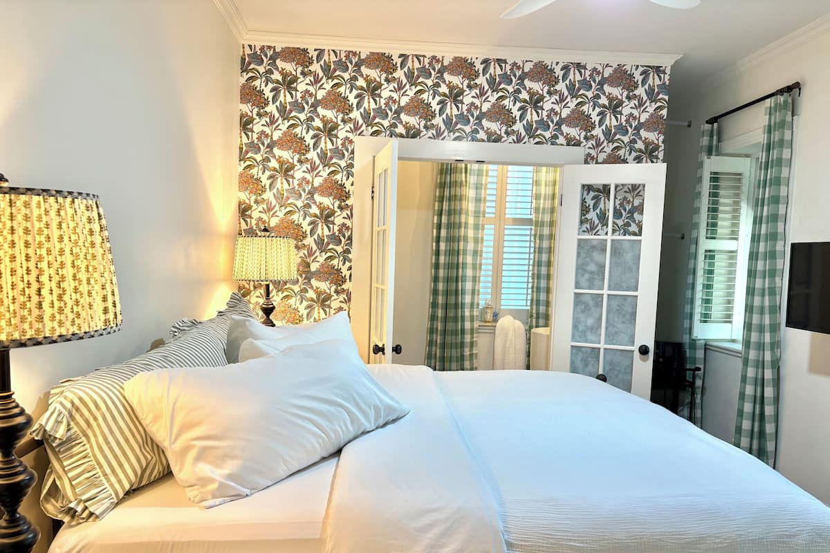 A hotel room with a bed, a lamp, a window with curtains, and a door leading to another room.