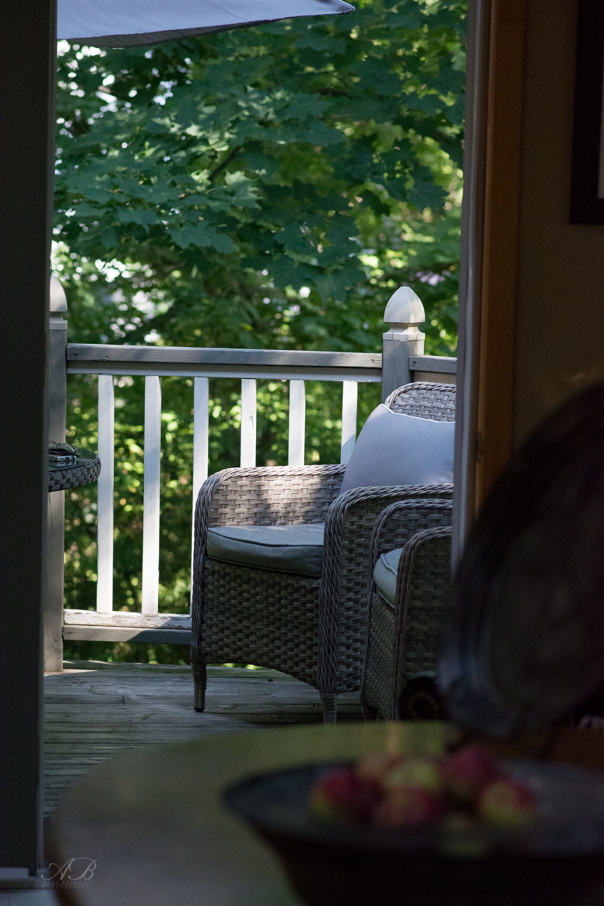 A wicker chair with a cushion on a wooden deck, a white railing, and a view of green foliage.