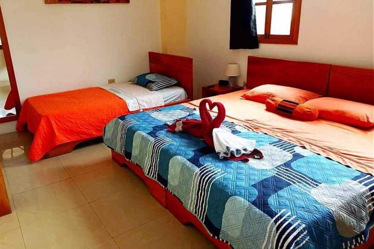 Une chambre d’hôtel avec deux lits, l’un recouvert d’une couverture rouge et l’autre d’une couverture à motifs bleus. Il y a un tableau au mur et une fenêtre avec un rideau.