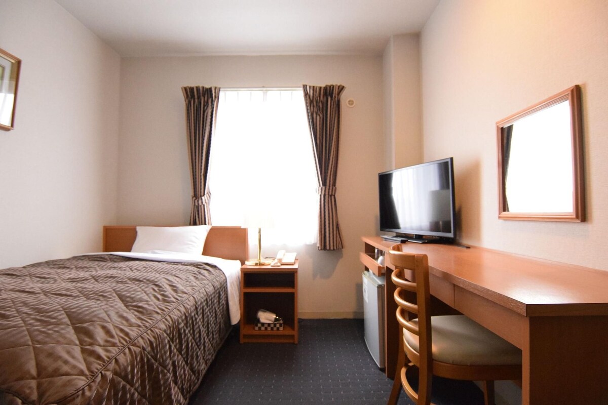 A hotel room with a bed, a desk with a television, a chair, a small shelf, and a window with curtains.