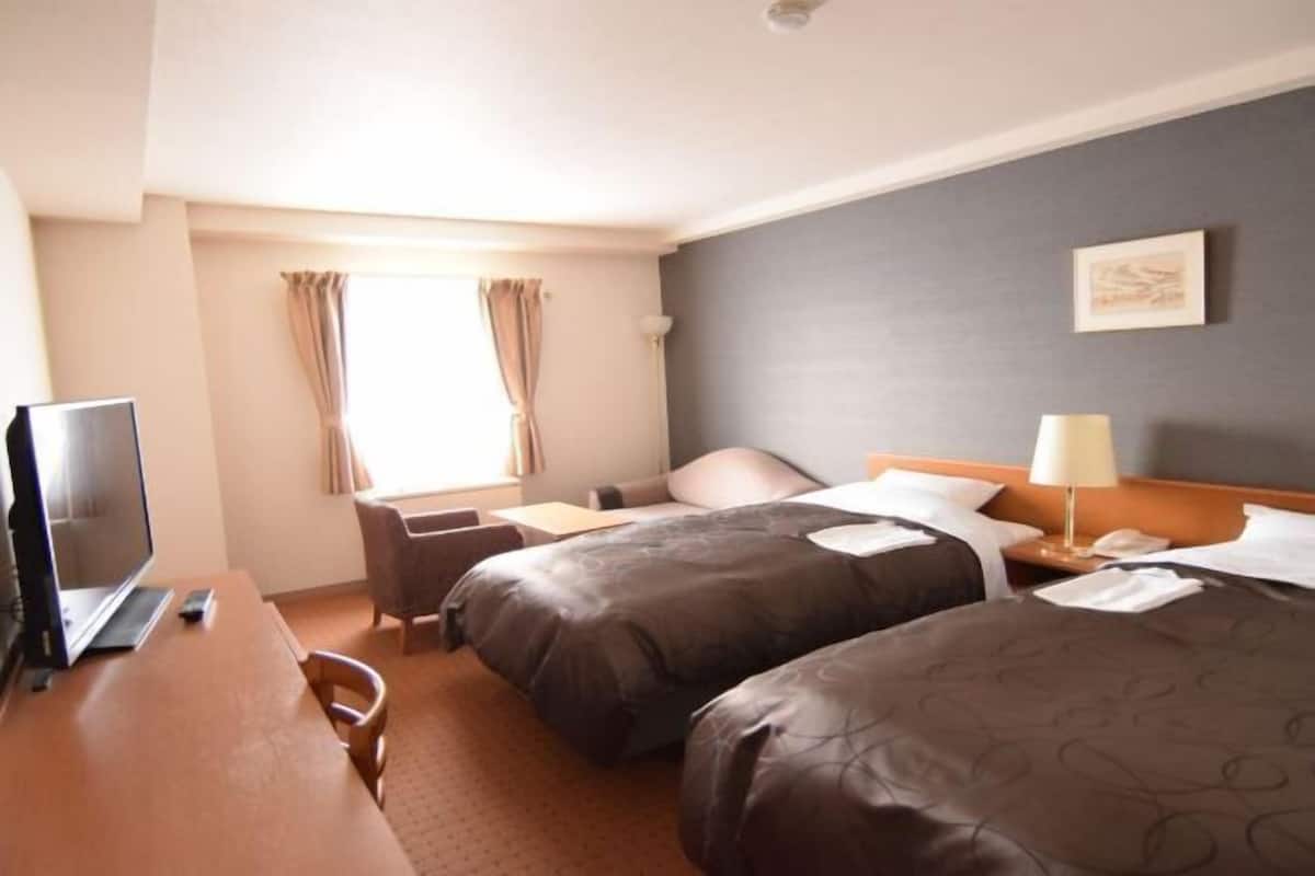 Ein Hotelzimmer mit zwei Betten, einem Schreibtisch mit einem Laptop, einem Stuhl, einer Schreibtischlampe und einem Fenster mit Vorhängen.