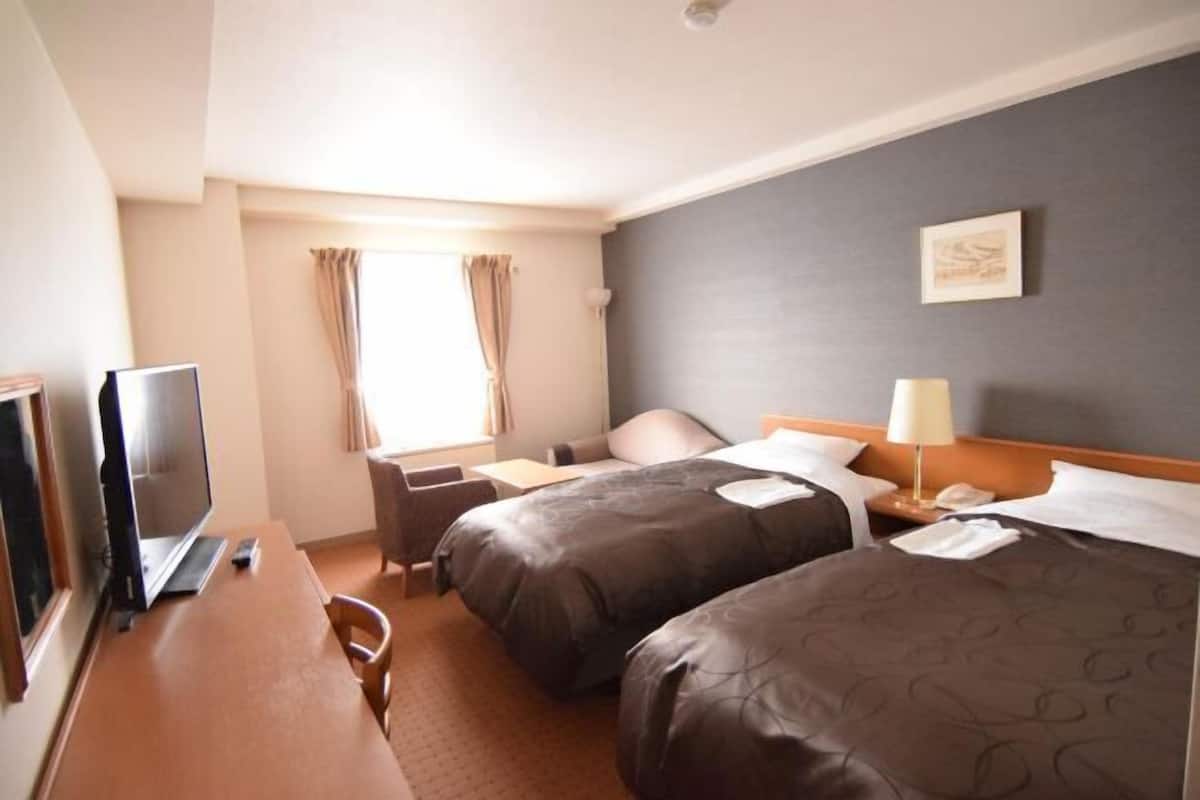 Ein Hotelzimmer mit zwei Betten, einem Schreibtisch mit Computer, einem Fernseher und einem Fenster mit Vorhängen.
