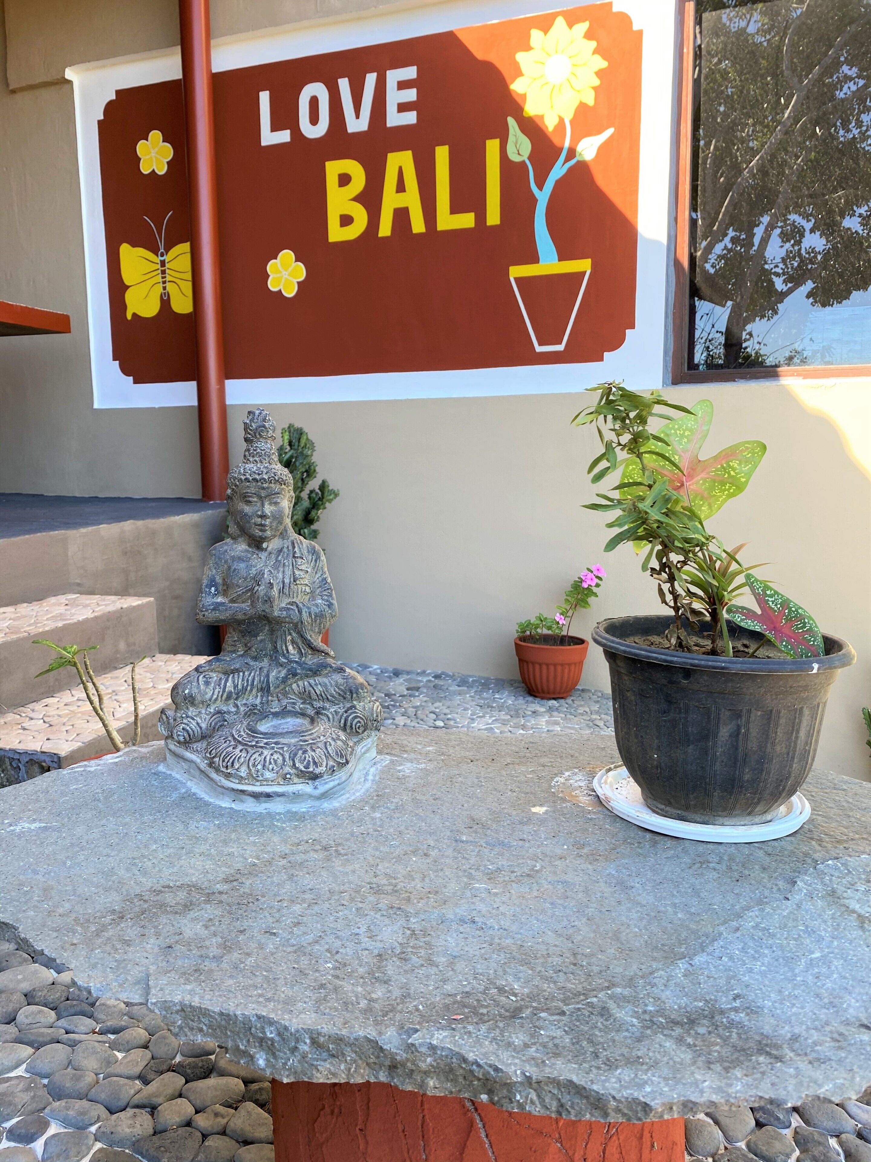 Eine Buddha-Statue aus Stein, ein Blumentopf mit Pflanze sowie ein Schild mit der Aufschrift 'LOVE BALI' und einer Blume.