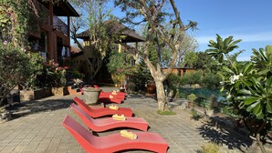 A patio with red lounge chairs, a large planter, and a pool in the background. - Villa Anjing 2 (Nusa Dua)
