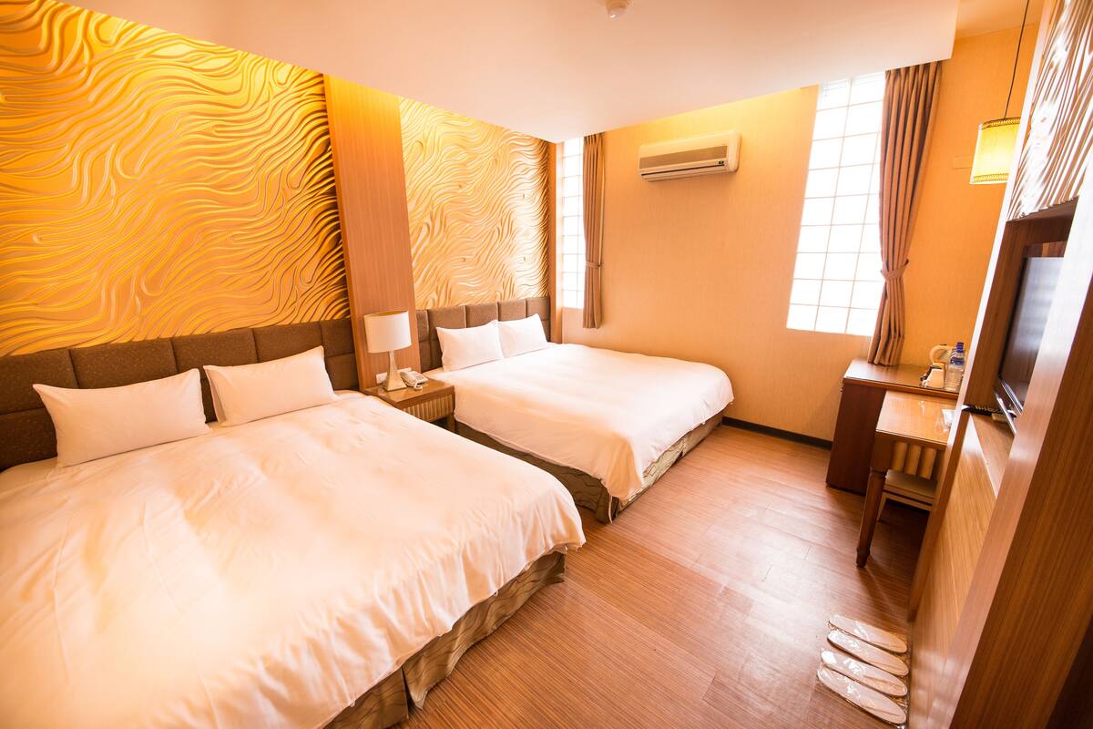 A hotel room with two beds, wooden flooring, a desk, a chair, an air conditioning unit, and a window with curtains.
