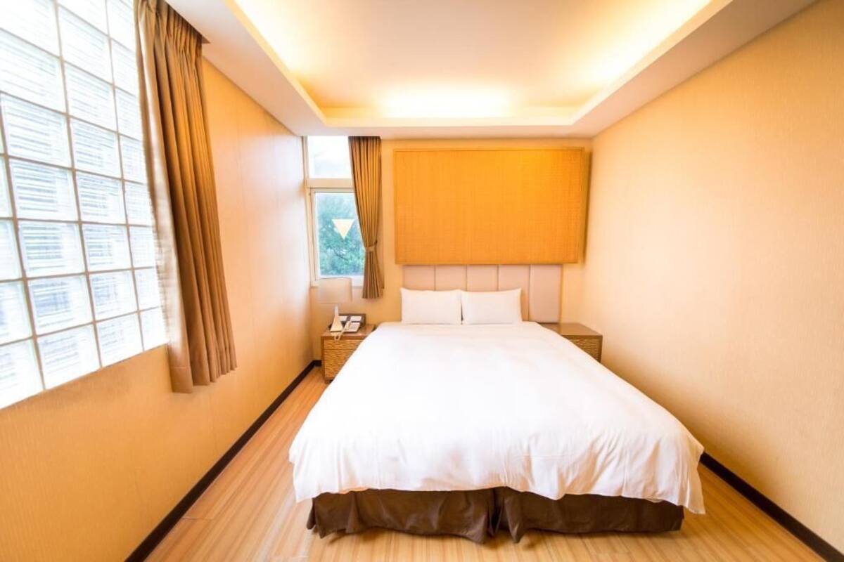 A hotel room with a large bed, a bedside table with a lamp, a window with curtains, and a headboard.