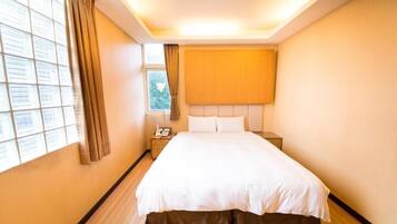 Una habitación de hotel con una cama grande, una mesita de noche con lámpara, una ventana con cortinas y un cabecero.