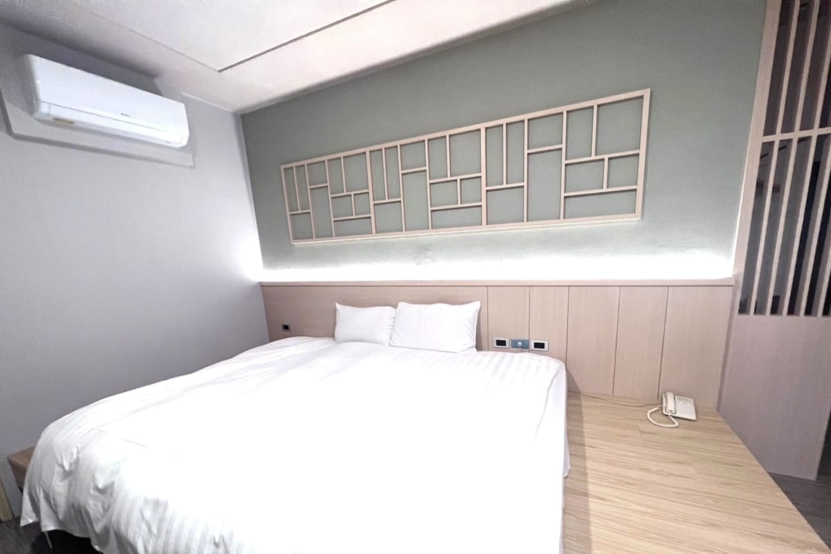A neatly made bed in a hotel room with a simple headboard, a wall-mounted air conditioner, and a telephone on the nightstand.