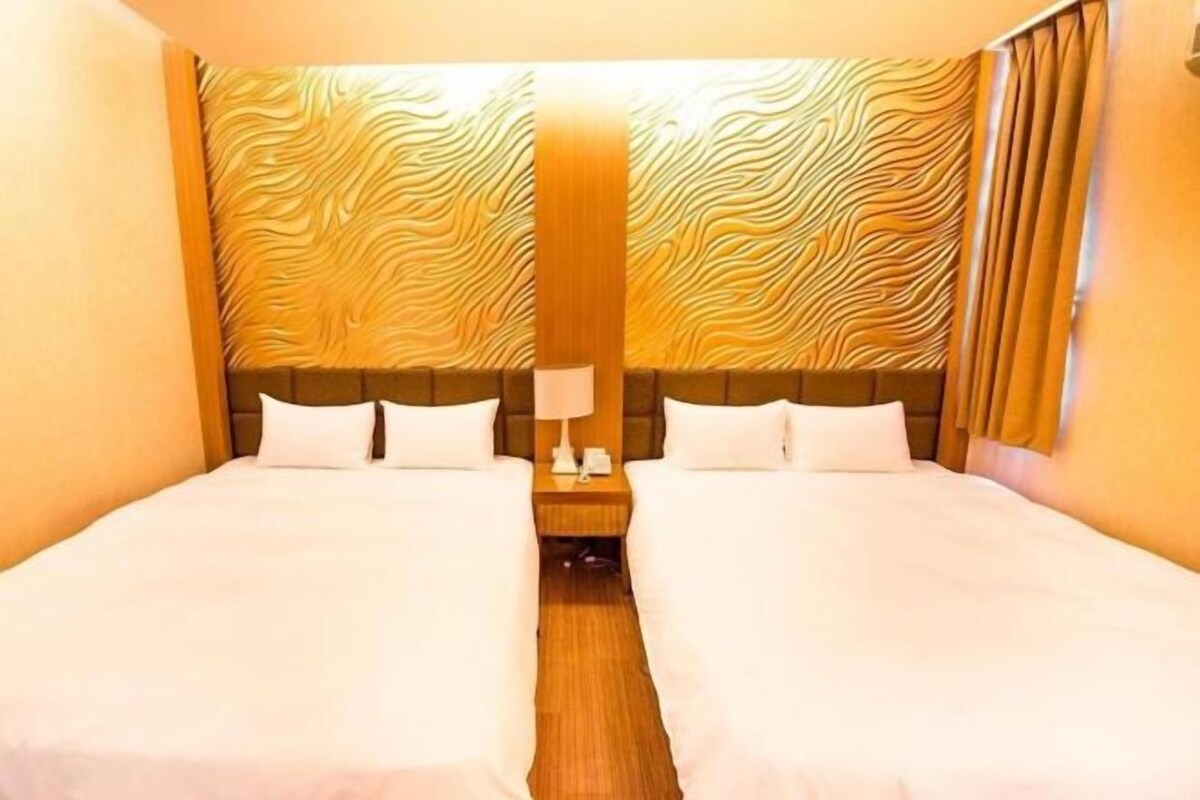 Una habitación de hotel con dos camas, cabecero de madera, mesita de noche con lámpara y pared con diseño.