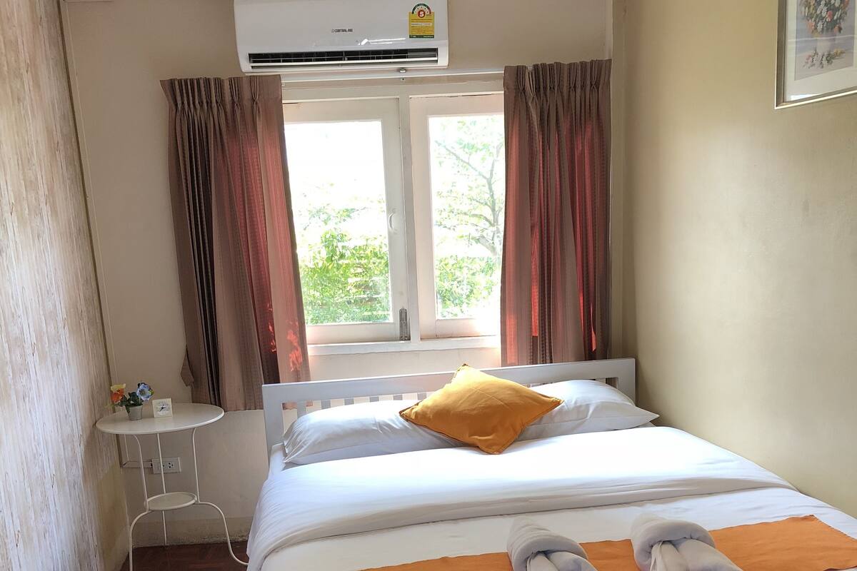 A hotel room with a bed, a small table, an air conditioning unit, a window with curtains, and a view of greenery outside.