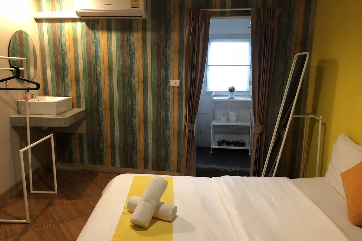 A hotel room with a bed, a bathroom visible through a door, a towel on the bed, and a shelf with toiletries.