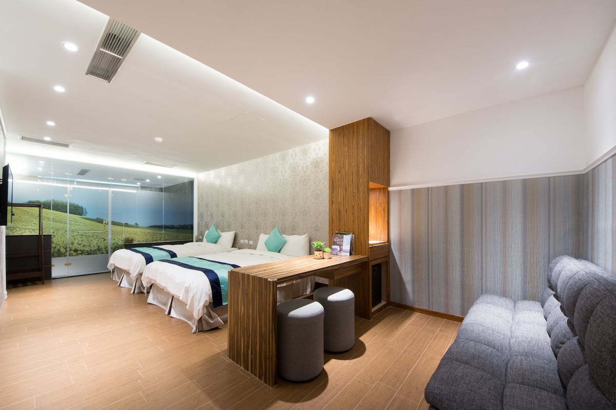 A modern hotel room with a large bed, a desk, two ottomans, a sofa, and a large window with a scenic view.
