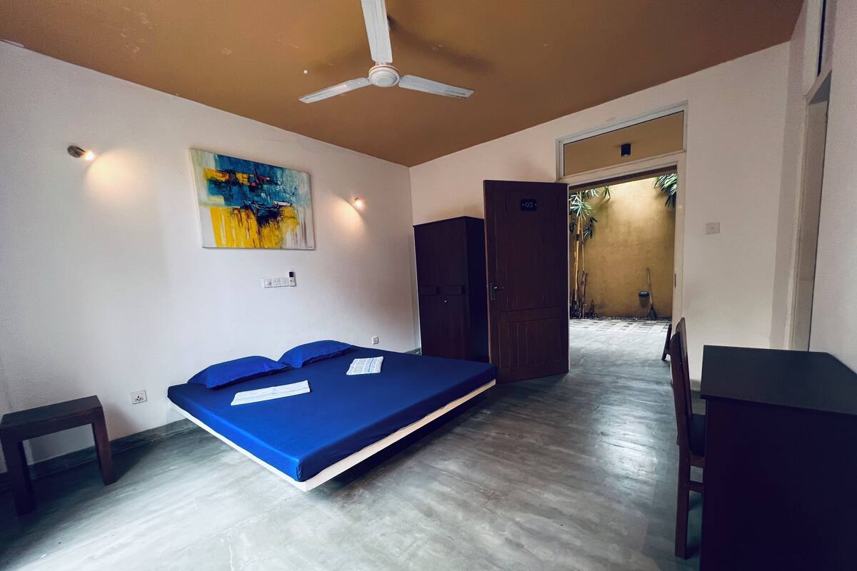 A room with a bed, a desk, a chair, a ceiling fan, and a painting on the wall.
