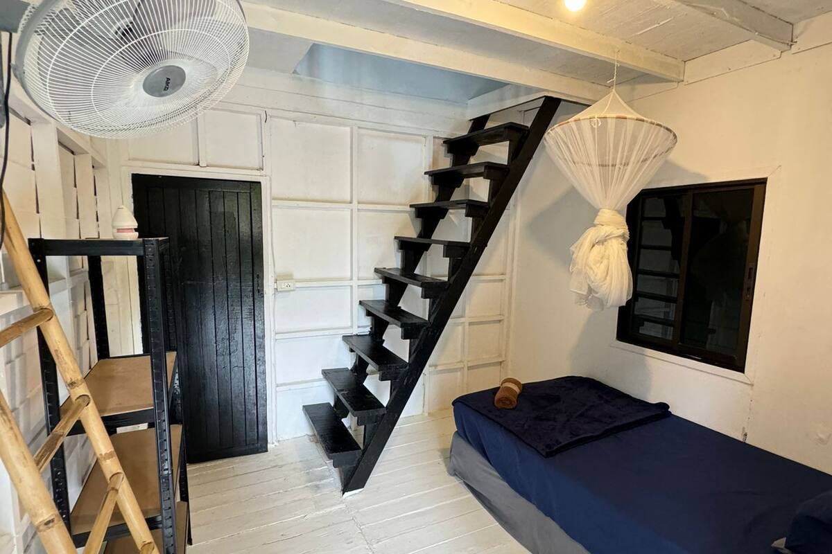 A room with a white wooden floor, a black staircase, a bed with a blue mattress, a fan, and a window with a white curtain.