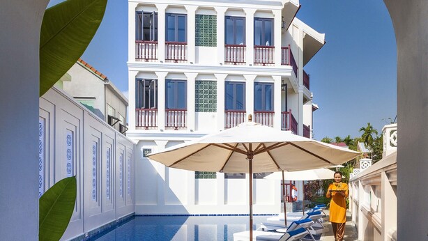 Una piscina con sillas de descanso y una sombrilla grande, un edificio con balcones, y un cielo azul despejado.