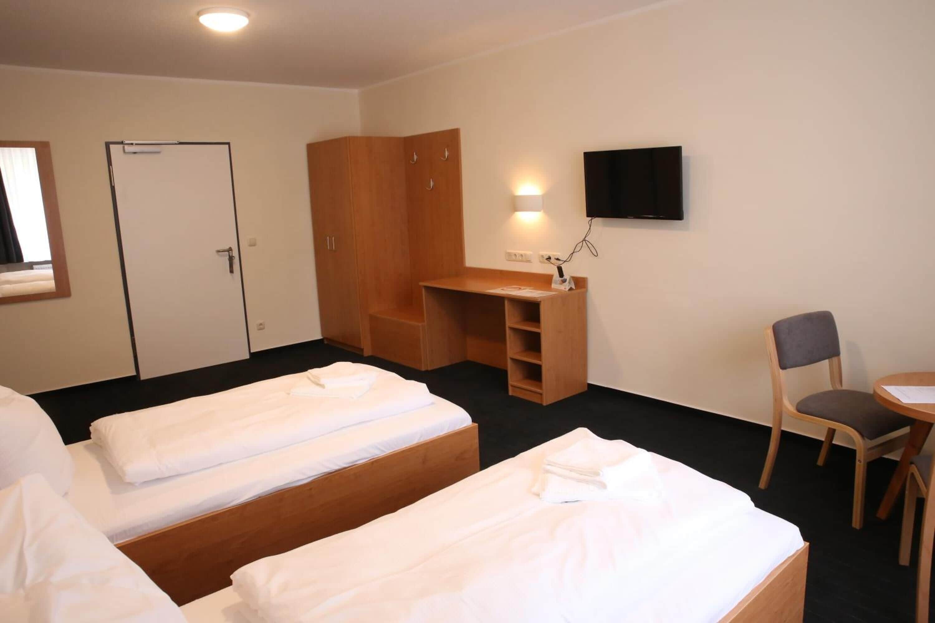 Quarto de hotel com duas camas de solteiro, uma secretária de madeira, uma cadeira, televisão e guarda-roupa.