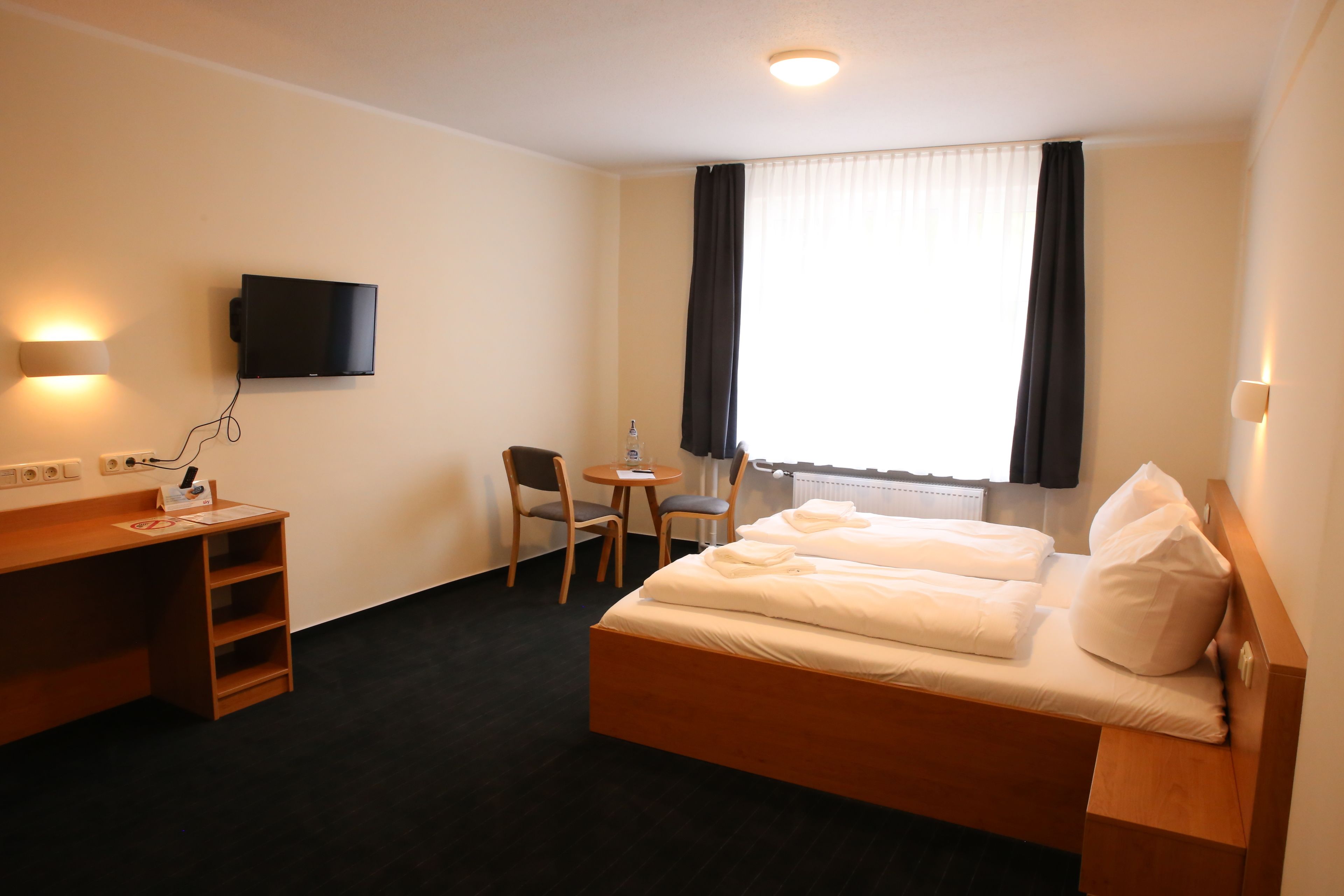 A hotel room with a bed, a desk with a chair, a TV mounted on the wall, and a window with curtains.