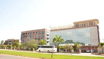 A modern hotel with a glass facade, a bus parked in front, and palm trees.