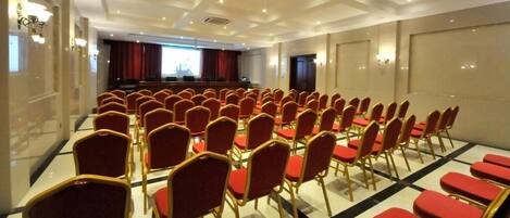 A conference room with rows of red chairs and a stage with a projector.