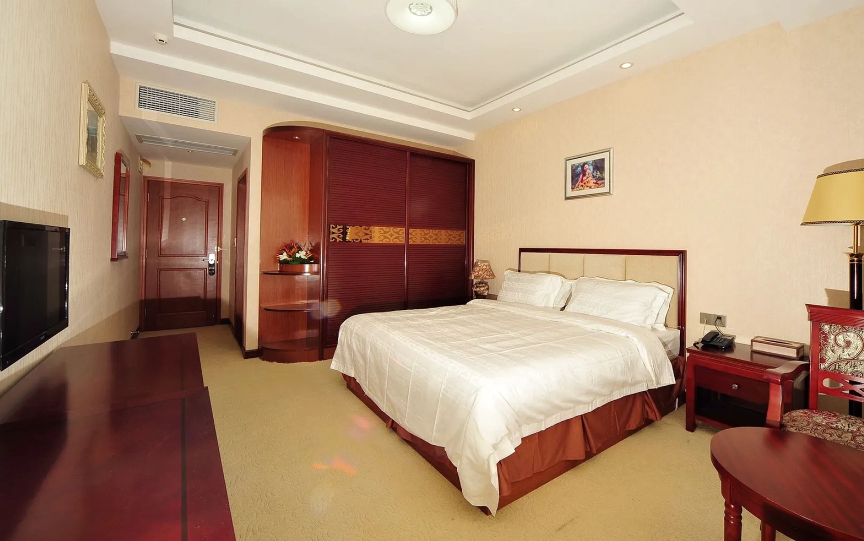 A hotel room with a large bed, a desk, a TV, a wardrobe, and a bedside table with a lamp.