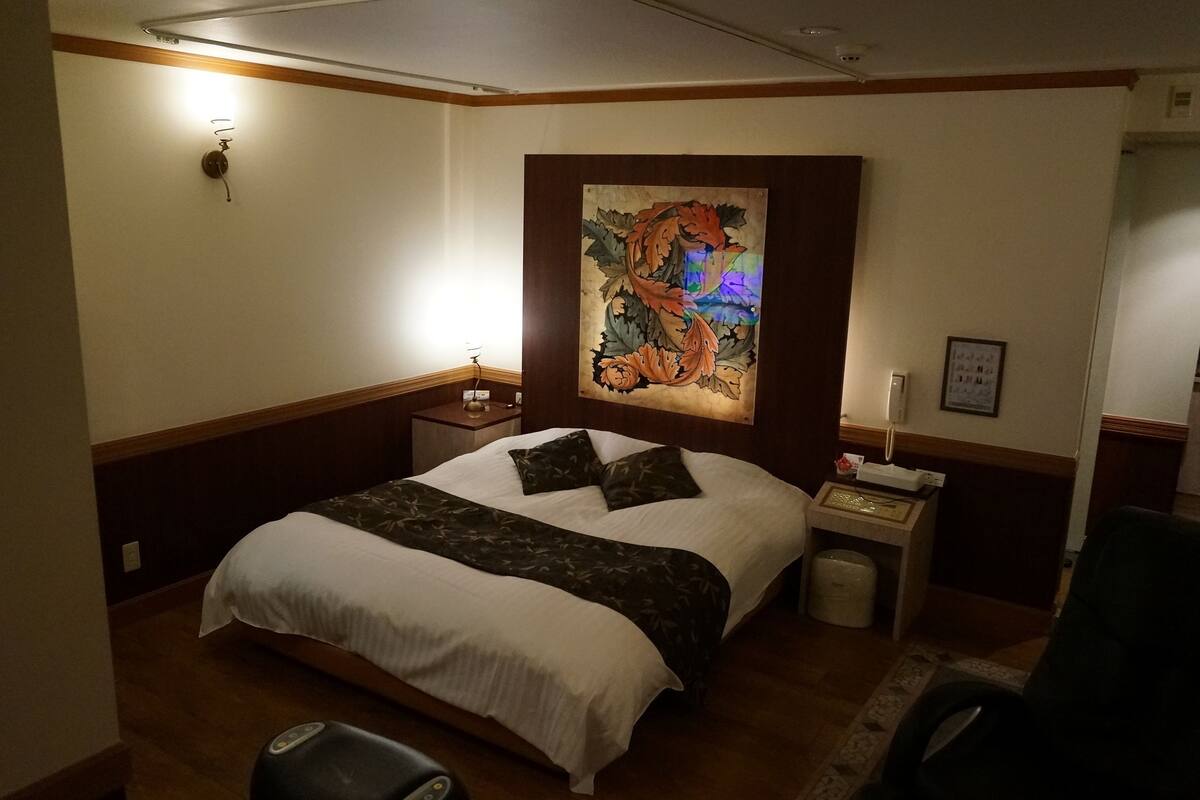 Una habitación de hotel con cama, mesita de noche, lámpara, sillón y un cuadro en la pared.