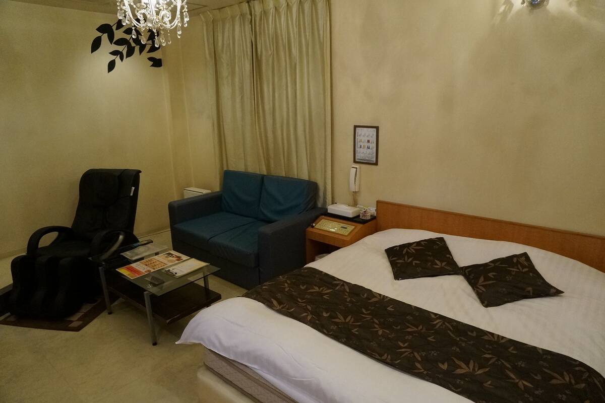 Habitación de hotel con una cama, un sillón azul, una mesita con una revista, una silla de masaje negra y un teléfono en la mesita de noche.
