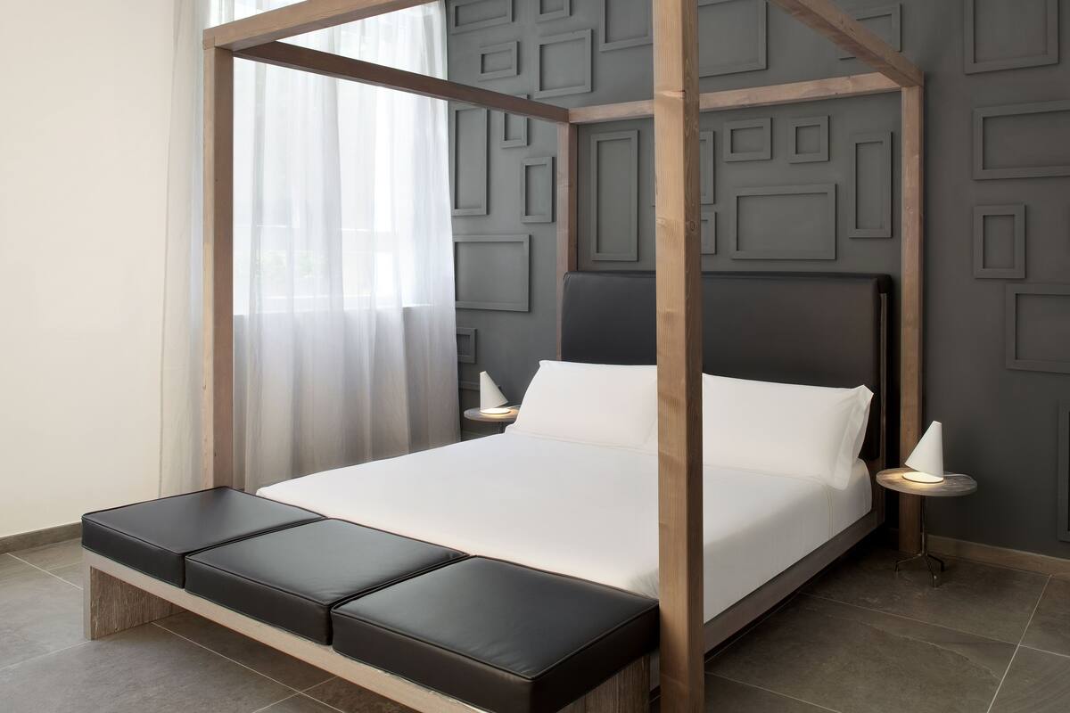 A modern bedroom with a wooden canopy bed, a bench at the foot, a bedside table with a lamp, and a wall with geometric paneling.