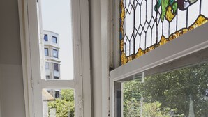 A room with a white chair, a small wooden table, and a window with a view of buildings and trees. - Urban House Hostel (San Sebastián)