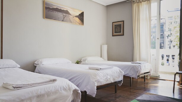 A hotel room with three single beds, a large window with curtains, a painting on the wall, and a view of the city. - Urban House Hostel (San Sebastián)