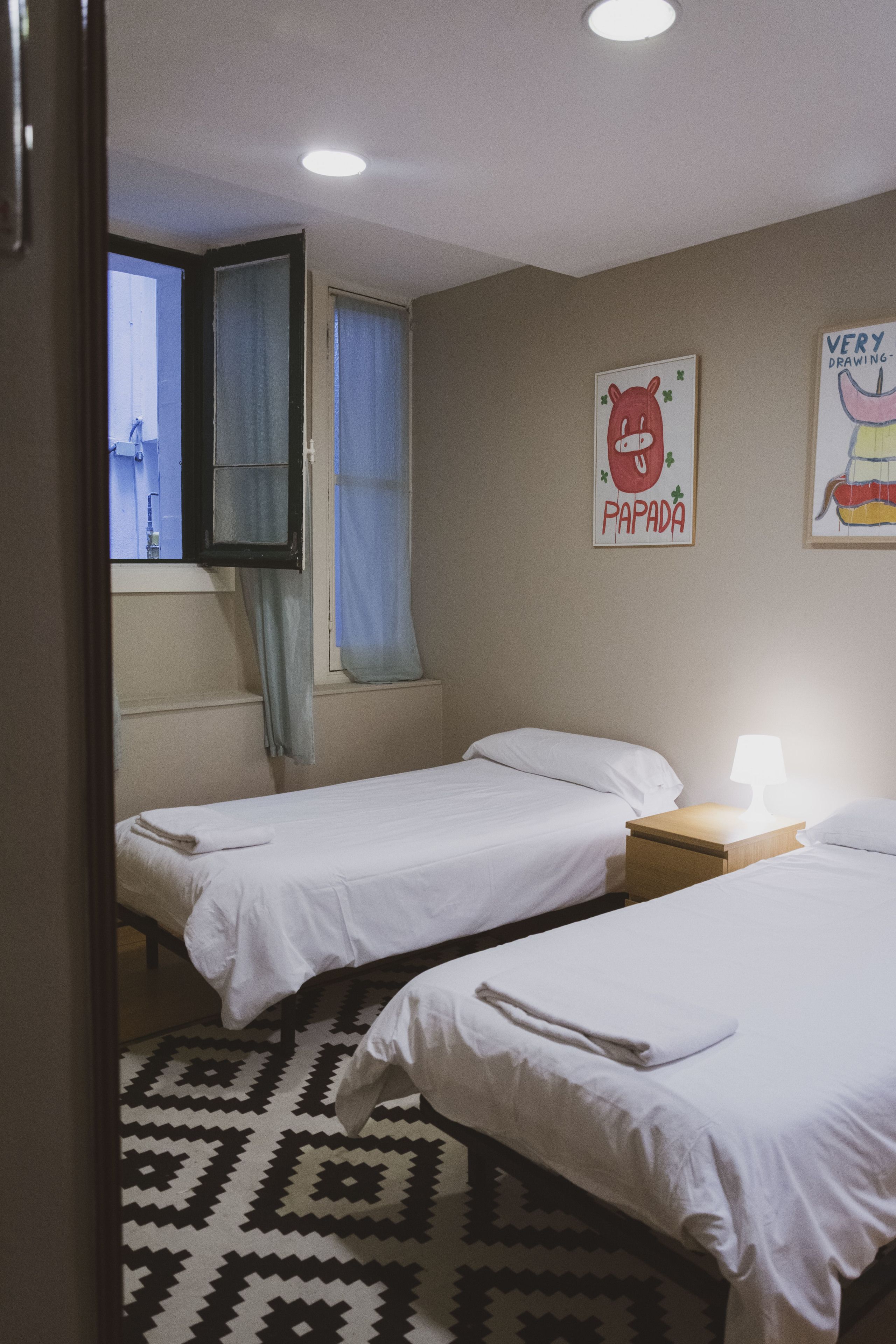 A bedroom with two beds, a window with curtains, a wall with two posters, and a patterned rug.